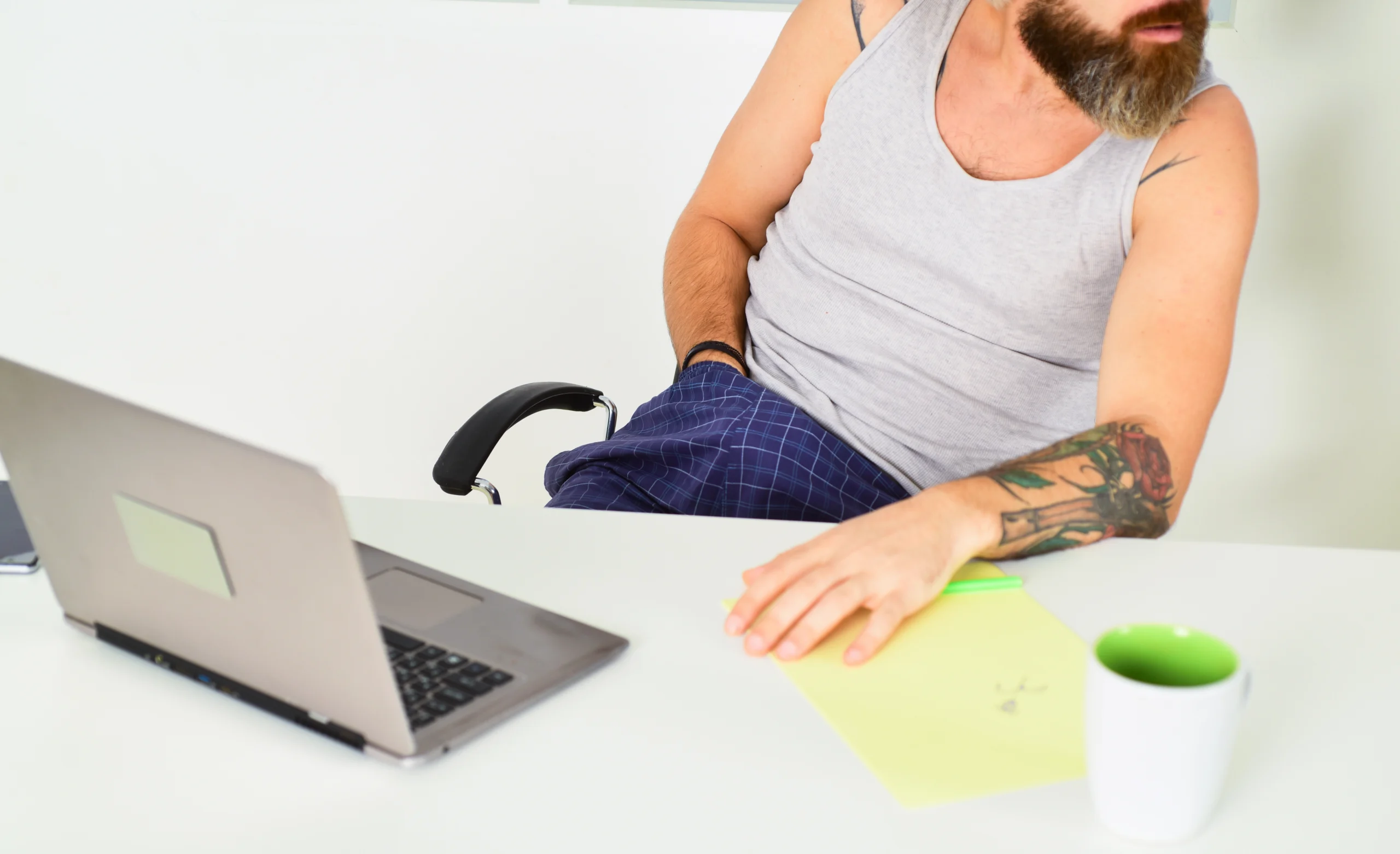 Masturbation. A man in a tank top and plaid pajama pants sits at a desk with a laptop, notepad, and green mug. His hand rests on the notepad and he appears to be looking away.