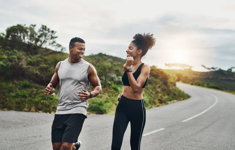 ED. Two people jogging together on a curved road outdoors, surrounded by greenery, with the sun low in the sky.
