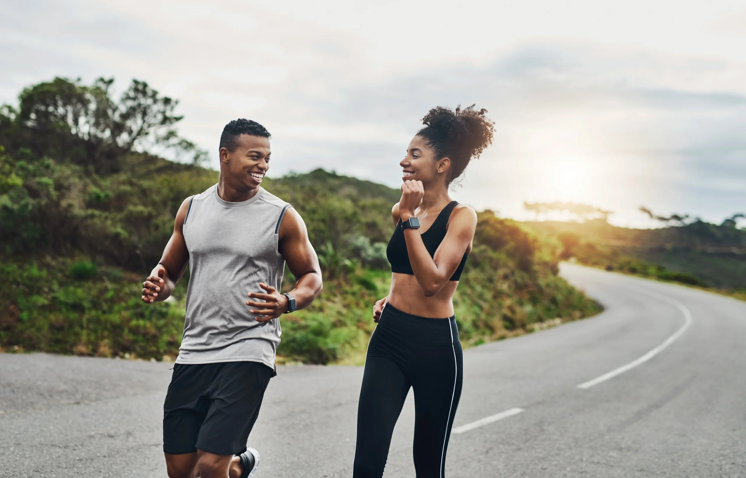 ED. Two people jogging together on a curved road outdoors, surrounded by greenery, with the sun low in the sky.