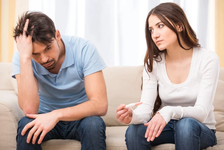 male infertility. A woman holding a pregnancy test sits next to a man with his hand on his head; both appear serious and concerned on a couch.