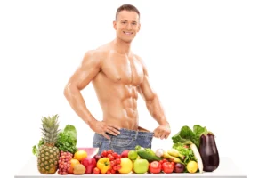 Shirtless man smiling and holding out the waistband of loose jeans, standing behind a table filled with various fresh fruits and vegetables.