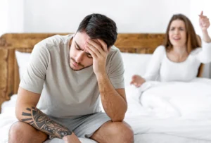 premature ejaculation. A man sits on the edge of a bed holding his head, looking distressed, while a woman in the background appears to be arguing or gesturing from the bed.