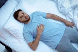 erectile dysfunction. A man in a blue shirt lies on his back in bed, looking up with one hand on his chest and the other resting by his side.