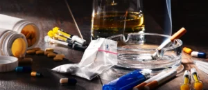 erectile dysfunction. Various drugs, pills, syringes, a plastic bag with white powder, a cigarette in an ashtray, a lighter, and a glass of alcohol are displayed on a dark wooden surface.