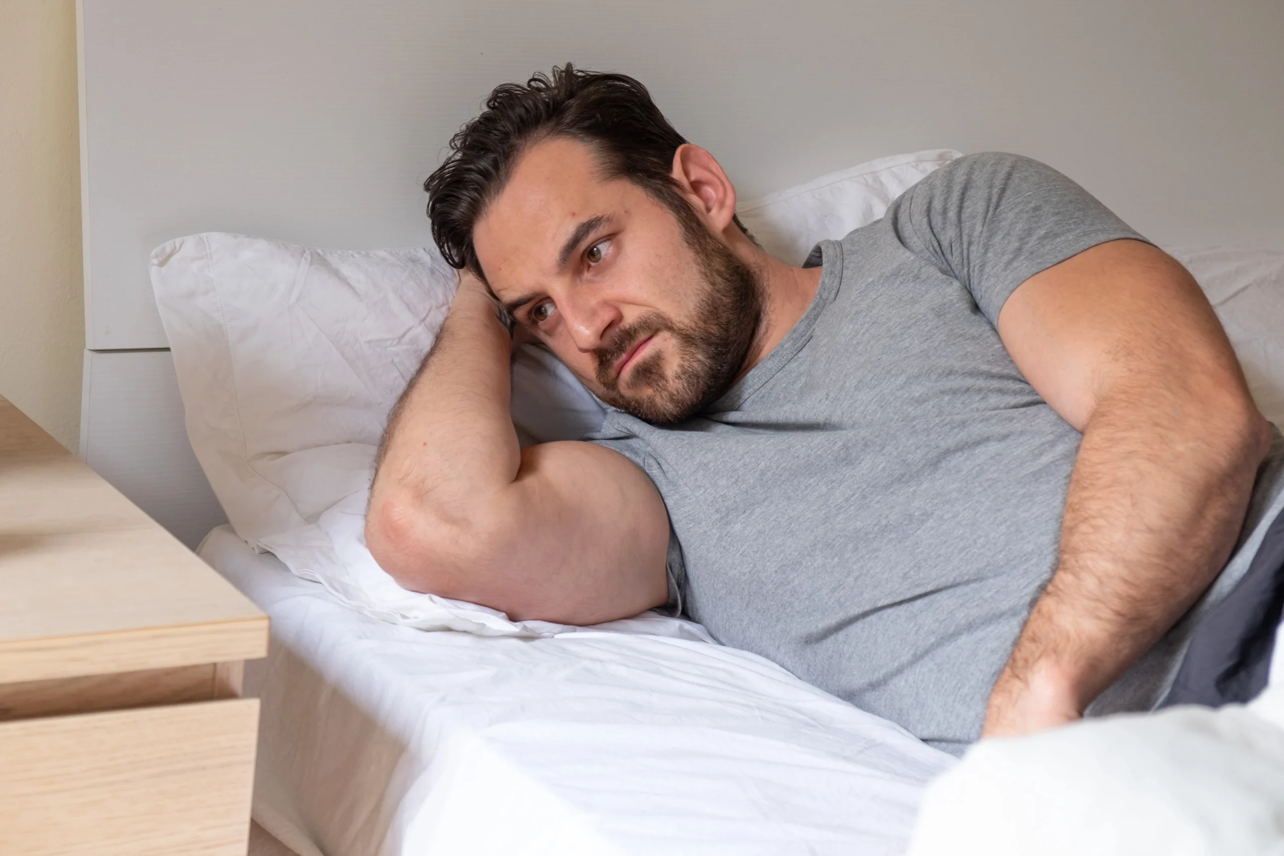 Low testosterone. A man with a beard lies on his side in bed, propped up on one elbow, looking thoughtful or concerned.