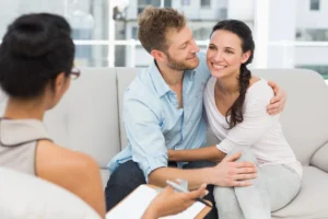 ED. A smiling couple sits close together on a couch during a counseling session, while a therapist with a clipboard listens and takes notes.