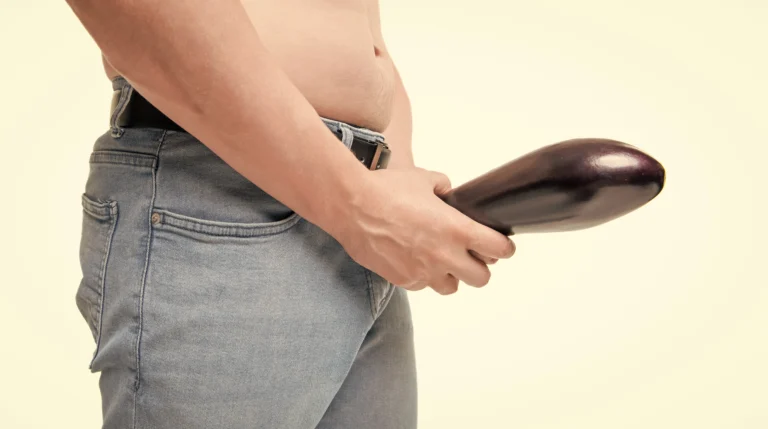 Masturbation. A person in jeans holds an eggplant at their groin area, partially exposing their torso against a neutral background.