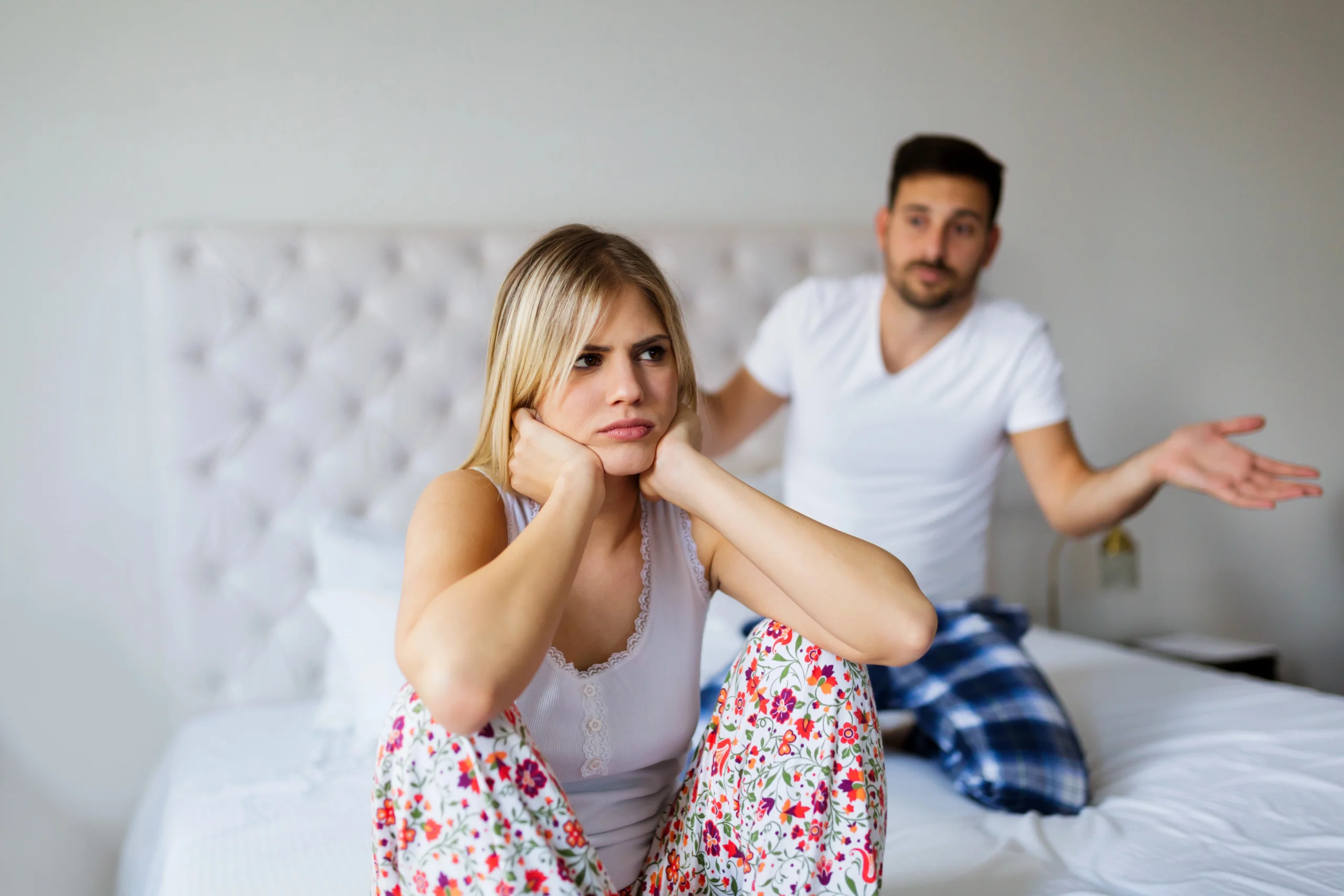 erectile dysfunction. A woman sits on a bed with a frustrated expression, while a man in the background gestures as if explaining or arguing.