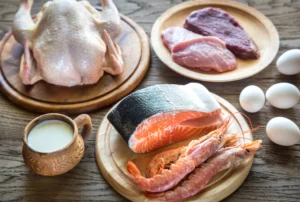 Natural foods. Various protein-rich foods including a whole raw chicken, steak, pork, salmon, shrimp, eggs, and a cup of milk arranged on a wooden table.