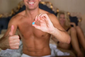 ED treatment. Shirtless man sitting on a bed holds a blue pill in one hand and gives a thumbs-up with the other, while a woman sits in the background.