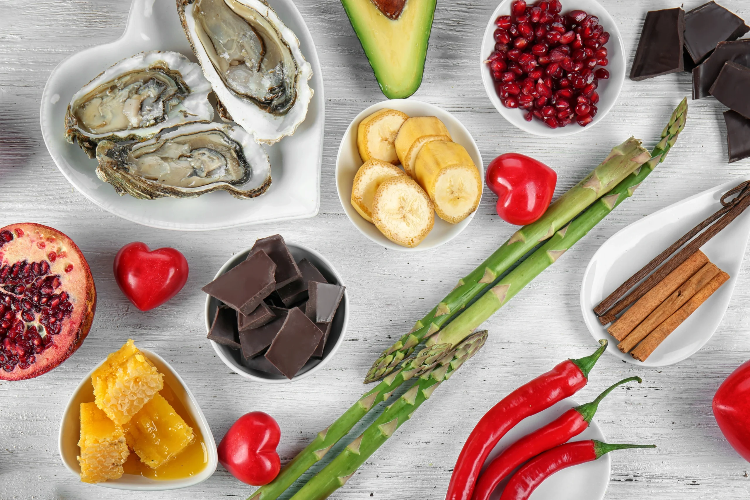 Natural foods. Various foods including oysters, avocado, pomegranate seeds, banana slices, chocolate, asparagus, honeycomb, chili peppers, and cinnamon sticks arranged on a white wooden surface.