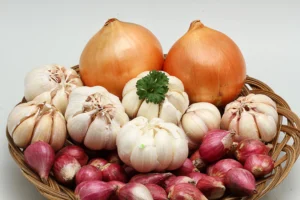 Natural foods. A basket containing two brown onions, several garlic bulbs, and small red shallots, with a sprig of parsley placed on top, against a plain white background.