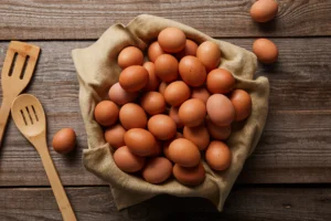 Natural foods. A cloth-lined bowl filled with brown eggs sits on a wooden surface, with two wooden spatulas and a few eggs scattered nearby.