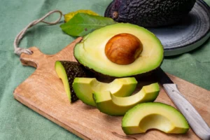 Natural foods. A halved avocado with pit, sliced pieces, and a knife sit on a wooden cutting board; a whole avocado is in the background on a plate.