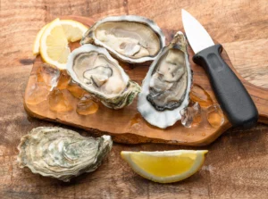 Natural foods. Three opened oysters on a wooden board with a shucking knife, ice cubes, and lemon wedges. One oyster shell is closed.