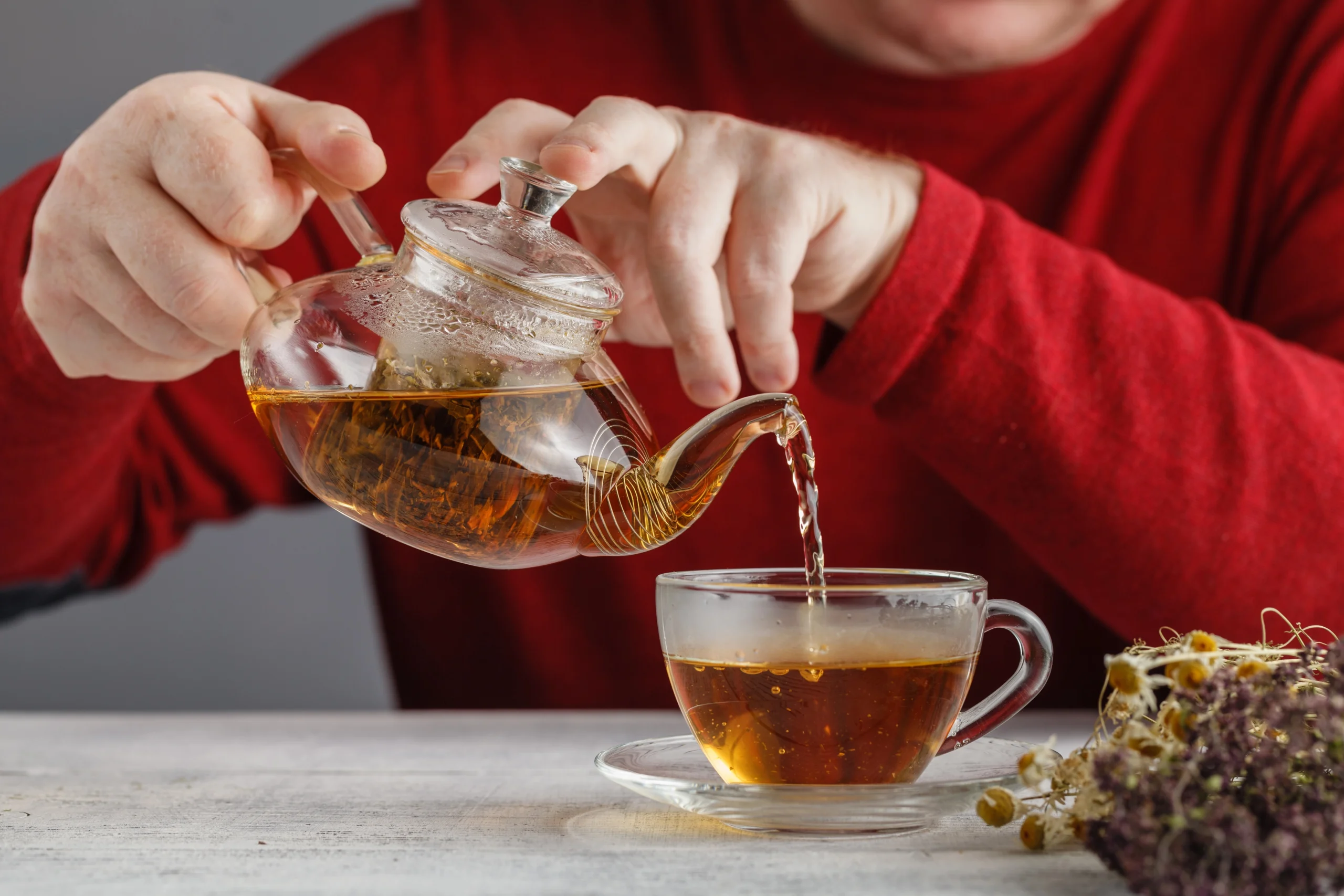 Erection tea. A person in a red sweater pours tea from a glass teapot into a clear teacup on a saucer, with dried flowers on the table.
