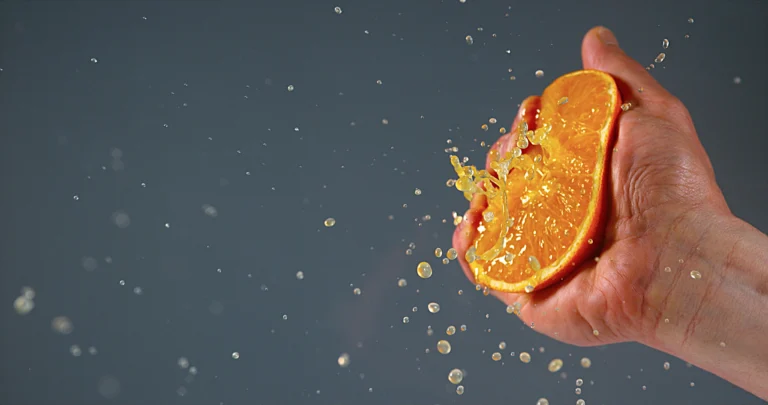 Squeeze technique. A hand squeezing a halved orange, with juice and droplets spraying outward against a plain gray background.
