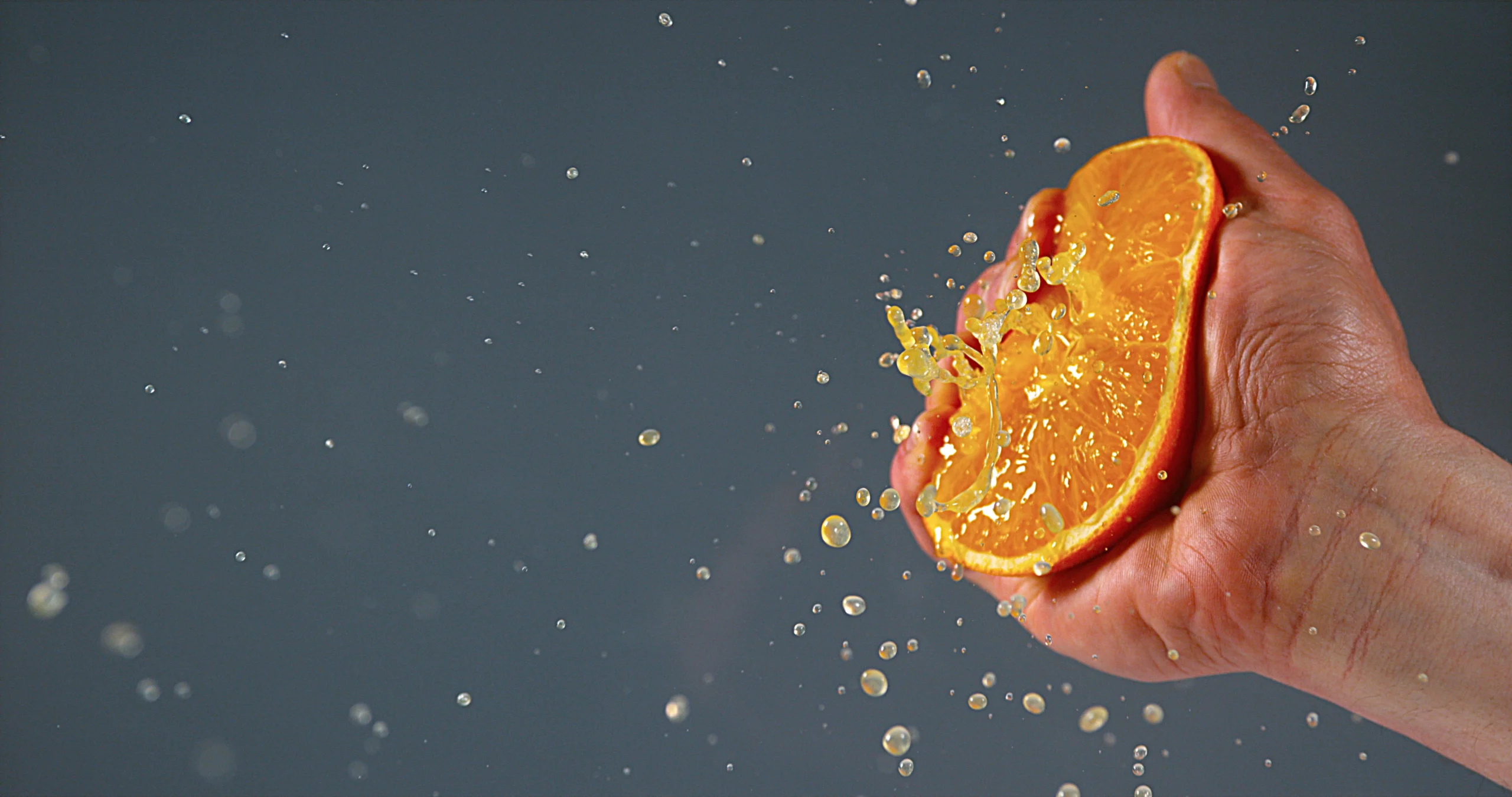 Squeeze technique. A hand squeezing a halved orange, with juice and droplets spraying outward against a plain gray background.