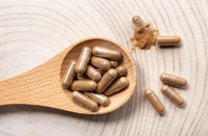 Ashwagandha. A wooden spoon filled with brown herbal capsules rests on a light wooden surface, with two capsules and some loose powder beside it.