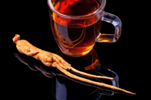 Erection tea. A glass cup of ginseng tea sits beside a whole ginseng root on a glossy black surface.