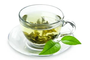 Erection tea. A glass cup of green tea with tea leaves submerged, placed on a glass saucer beside two fresh green tea leaves, all on a white background.