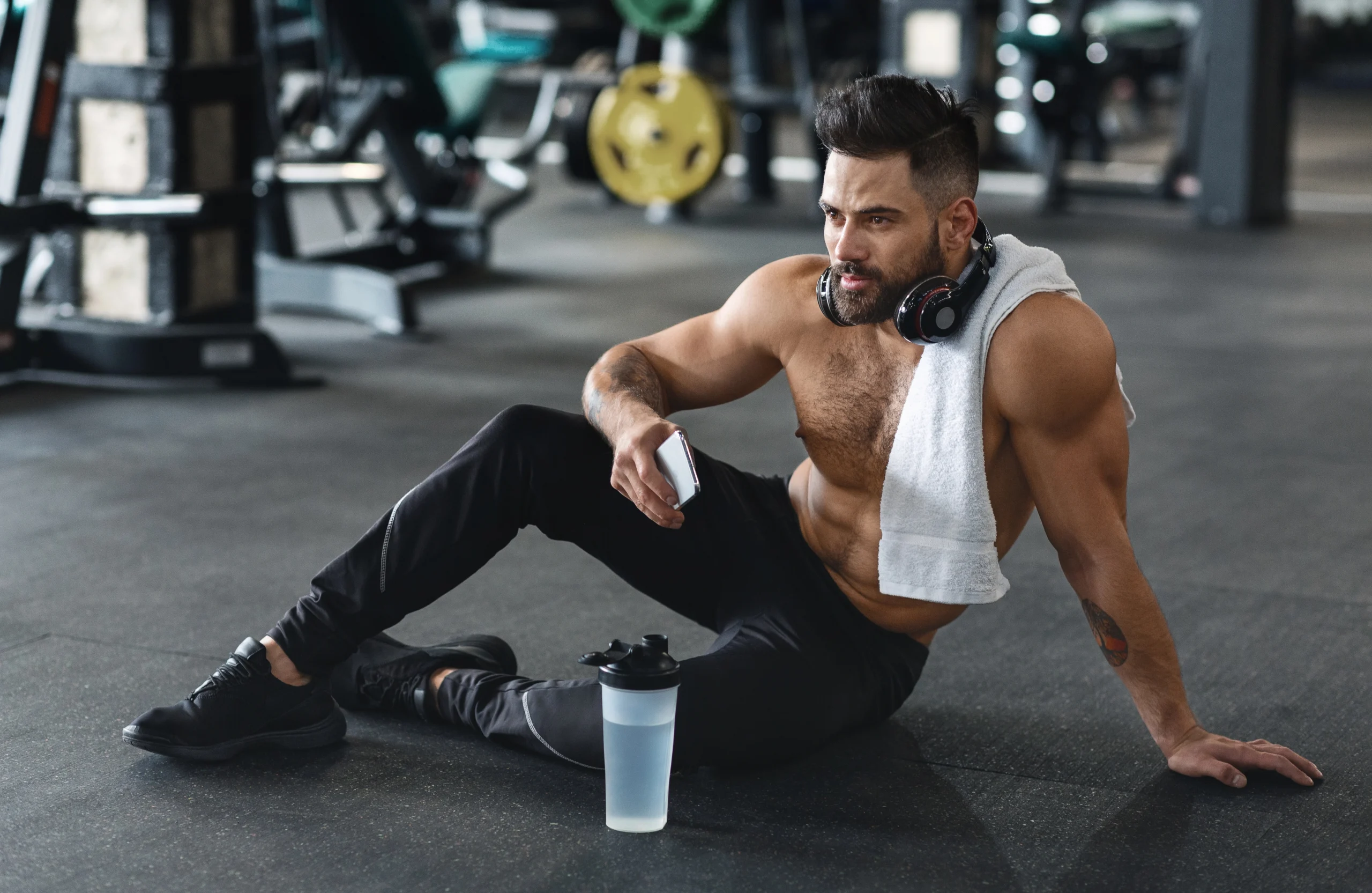 Kegel exercises. A shirtless man with a towel around his neck sits on a gym floor, wearing headphones and holding a phone. A protein shaker bottle is on the floor in front of him.