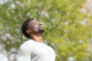 Sexual confidence. A man in a gray sweater stands outdoors with his eyes closed and head tilted slightly upward, appearing to take a deep breath. Trees with green foliage are blurred in the background.