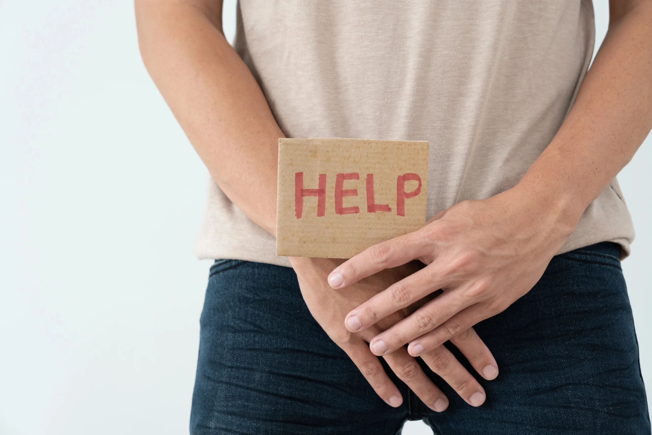 Premature ejaculation. A person stands with hands covering their crotch, holding a cardboard sign that says "HELP" in red letters.