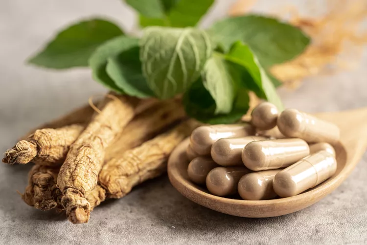 gingseng. A wooden spoon with herbal capsules sits beside ginseng roots and green leaves on a gray surface.