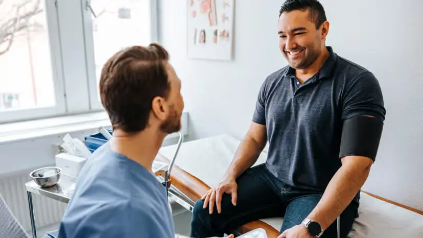 ginseng. A healthcare professional talks with a smiling man wearing a blood pressure cuff while seated on an exam table in a medical office.