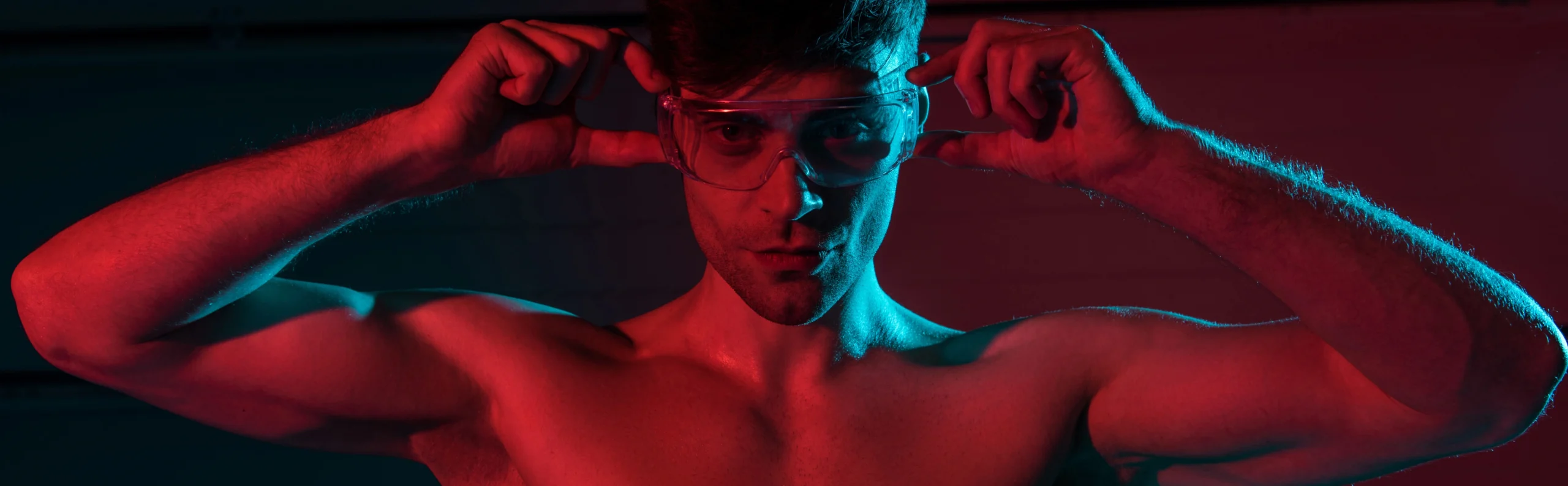 red light therapy. Shirtless man adjusts transparent safety glasses, illuminated by red and blue lighting, looking directly at the camera.