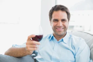 ED. Man in a light blue shirt sitting on a couch, smiling and holding a glass of red wine.