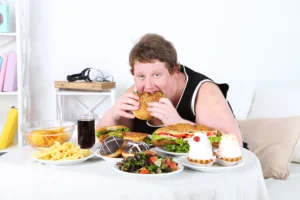 infertility. A person sits at a table eating a large burger, surrounded by fast food items, desserts, and a drink.