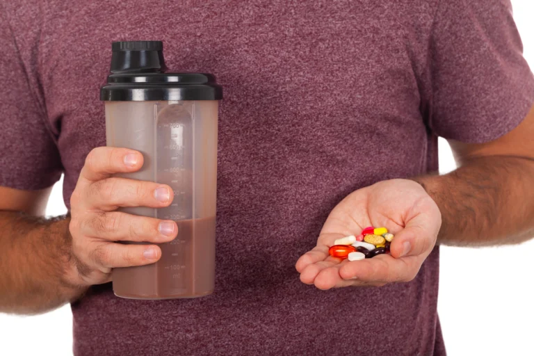 Testosterone. A person in a maroon shirt holds a protein shaker in one hand and several colorful pills in the other hand.