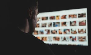 porn. A person sits in a dark room, looking at a computer monitor displaying a grid of blurred, colorful thumbnail images.