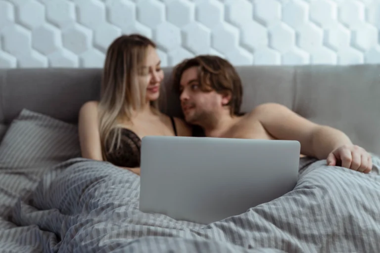 porn. A man and woman sit up in bed together under a striped gray blanket, looking at each other, with an open laptop in front of them.