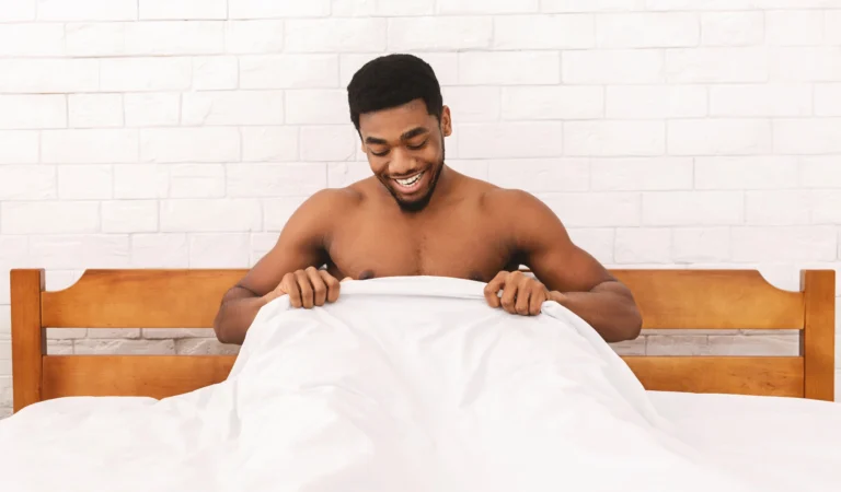 Testosterone. A man sits up in bed, holding a white blanket and smiling, with a wooden headboard and white brick wall in the background.