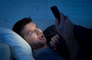 porn. Man lying in bed at night, holding a smartphone and looking at the screen with a surprised expression, illuminated by the phone's light.